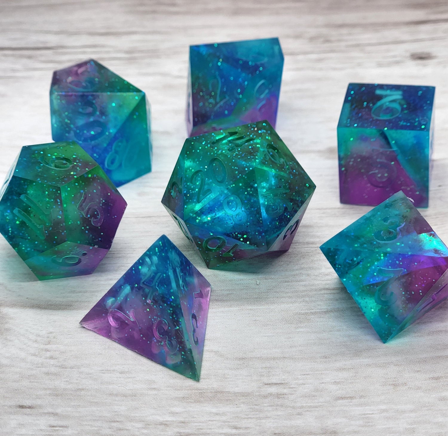 A set of 7 polyhedral dice that with a purple, blue, and teal galaxy look.